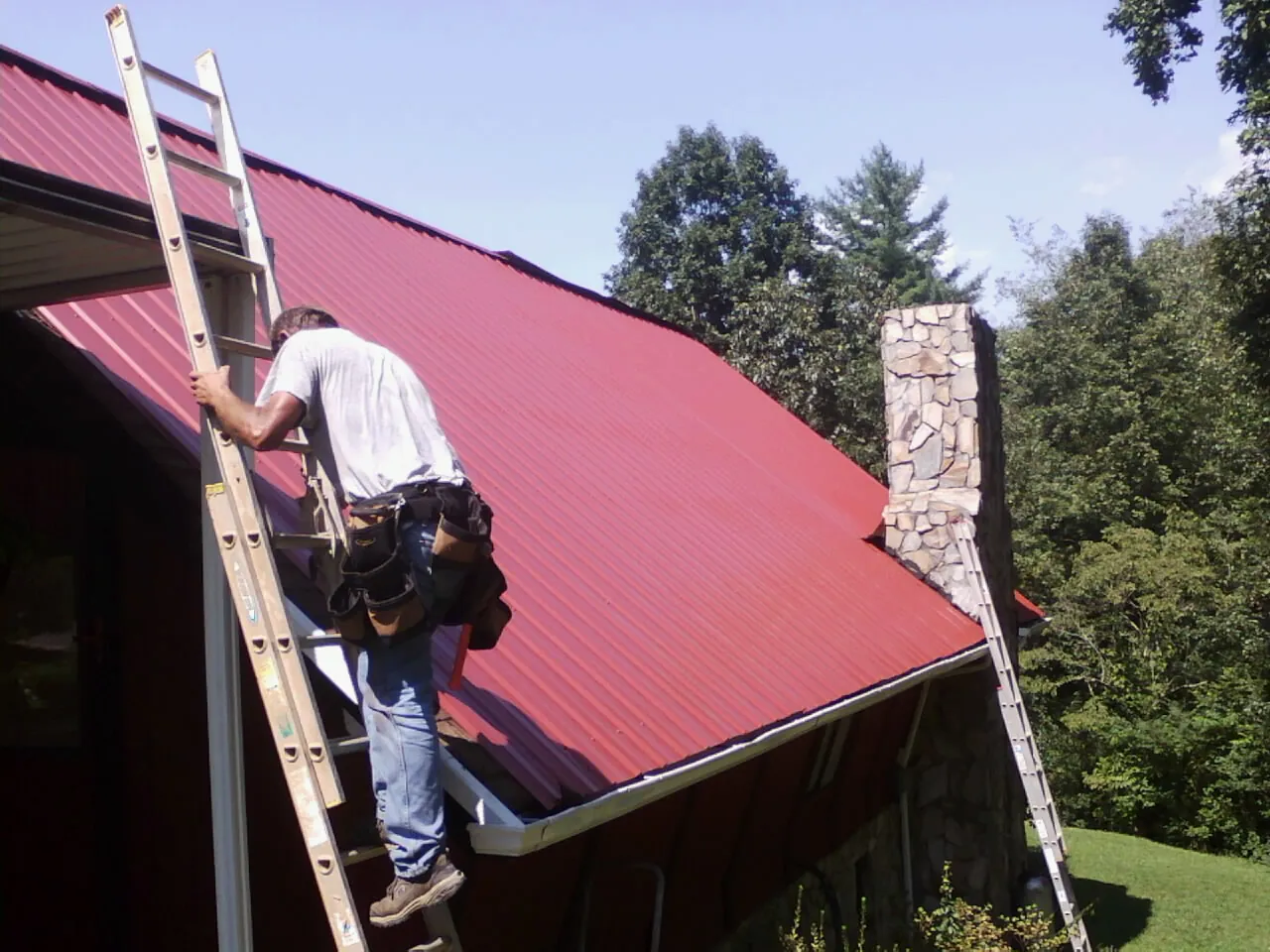 Roofer on a ladder beside a red metal roof and stone chimney — Soffit & Fascia Repair by Roofing Brothers in Rocklin